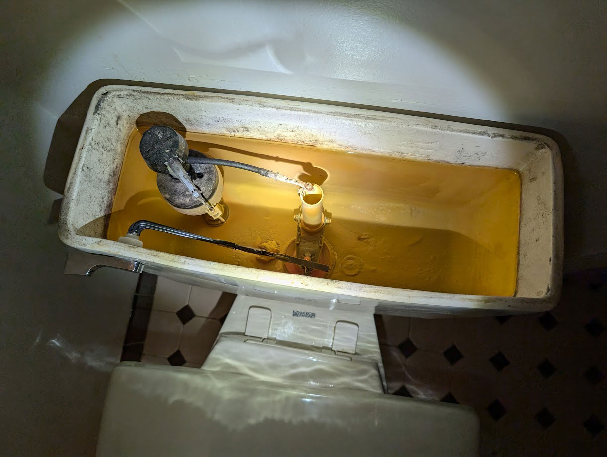 An opened toilet tank with a flashlight shining inside, revealing the float mechanism, fill valve, and water at the bottom. The tank interior appears aged with some rust and mineral deposits. The toilet base rests on a tiled floor.
