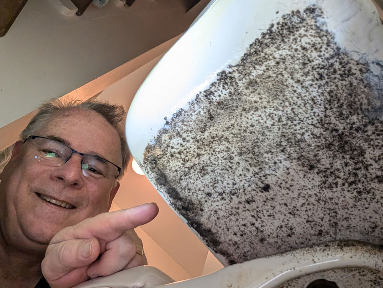 A person is pointing at the underside of a sink, which is covered in a black, mold-like substance. The person is smiling, wearing glasses, and is inside a room with a visible ceiling light.
