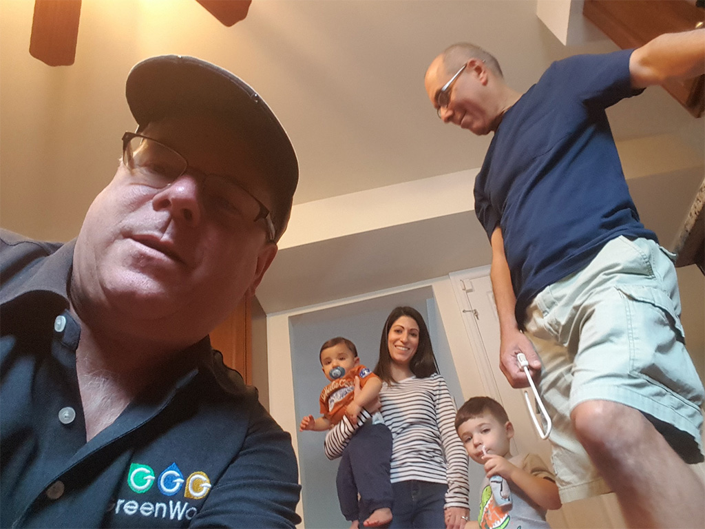Selfie of victor wearing a cap, with two people in the background, one of them holding children, in a home setting.