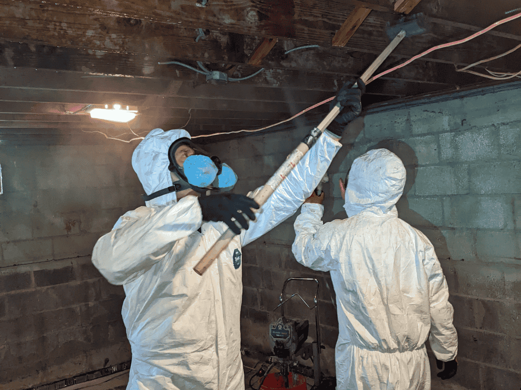 User-Inserted Two mold remediation professionals in protective suits treating contaminated crawl space ceiling and walls.