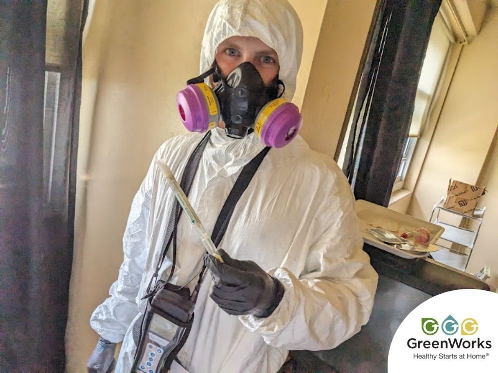 A mold expert in protective gear, including a respirator and full-body suit, conducting air quality testing with specialized equipment in an indoor setting.